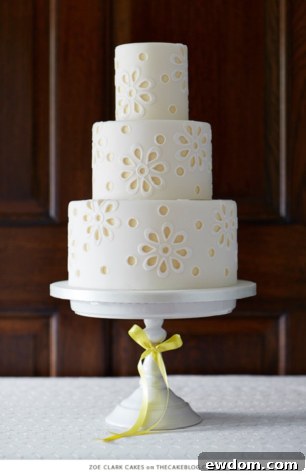 Elegant white cake featuring broderie anglaise inspired cut-outs revealing soft yellow accents and textured gumpaste appliqués by Zoe Clark Cakes.
