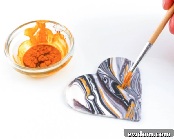 Applying edible gold luster dust mixed with clear alcohol to a marbled chocolate heart using a fine-tipped paintbrush, adding an elegant gilded detail.