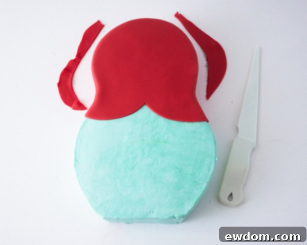 Matryoshka Magic Cake 12 Smoothing and trimming the red fondant headpiece on the Russian nesting doll cake for a clean finish. Cake by Cakegirls.