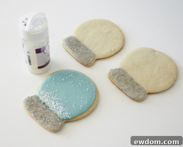 Decorating Snow Globe Cookies - Adding white nonpareils to the wet blue glaze to create a falling snow effect by Cakegirls for TheCakeBlog.com