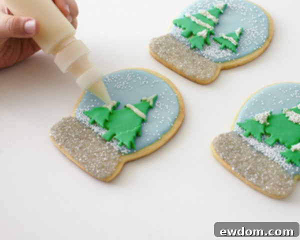 Decorating Snow Globe Cookies - Adding snow caps to the fondant Christmas trees with white glaze and nonpareils by Cakegirls for TheCakeBlog.com