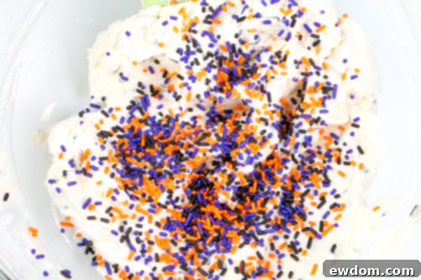 A mixing bowl with buttercream being combined with black, purple, and orange sprinkles, creating a festive base for the Halloween cake | by Erin Gardner for TheCakeBlog.com