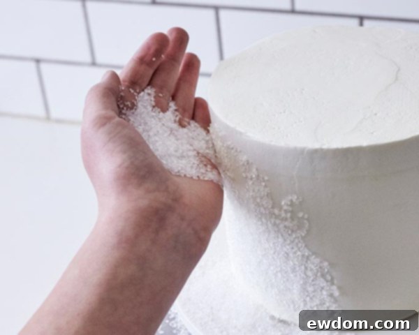 Enchanted Snowflake Cake 12 Applying white sugar crystals to the side of a frosted cake by hand